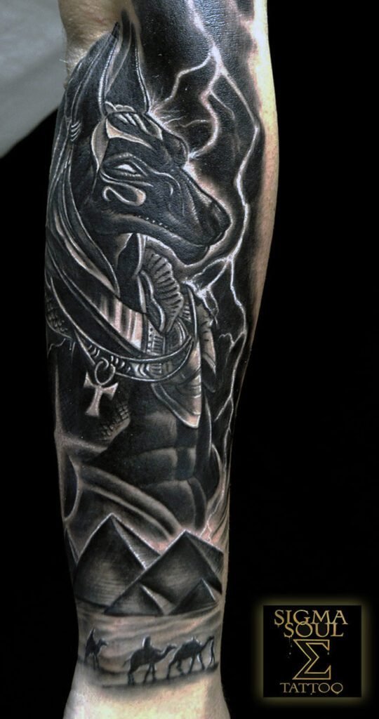 El guardián del umbral. Anubis, señor de las sombras y protector de las almas, revive en esta pieza donde mito y técnica convergen. Su mirada pesa, juzga y transmite autoridad. Representa el poder interior que entiende el equilibrio entre materia y espíritu. El diseño combina relieves metálicos, destellos y energía contenida. La iluminación resalta cada textura del collar y el manto, generando sensación de vida. Las pirámides y las caravanas completan la escena: el desierto como símbolo de eternidad. La composición proyecta fuerza, propósito y calma. Una obra pensada para atraer la mirada y provocar deseo. ¿Qué energía quieres despertar en tu piel? Descubre más en sigmasoultattoo.com o escríbenos por WhatsApp al 640 119 066.